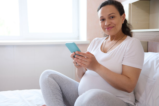 快乐的孕妇对着镜头微笑，使用智能手机，查看移动应用程序，看视频，计算婴儿的动作图片下载