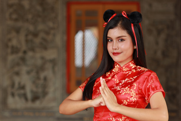 美丽的年轻亚洲女子，穿着红色的中国服装，传统的旗袍，她站在古老的墙前，做着祝贺的手势，脸上带着幸福的笑容。图片下载