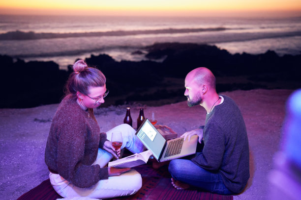 一对高加索数字游牧民夫妇悠闲地坐在沙滩上，喝着啤酒，在日落时分用笔记本电脑工作图片下载