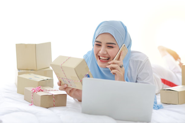 美丽和年轻的亚洲穆斯林妇女穿着睡衣，有吸引力的样子，躺在床上的电脑和在线包裹盒交付。创业中小企业自由职业女孩用手机工作图片下载