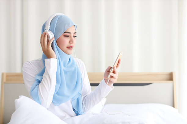 美丽的亚洲女性肖像，穿着睡衣，坐在床上，用手机看网络故事。年轻可爱的女孩戴着头巾，用智能手机听音乐图片下载