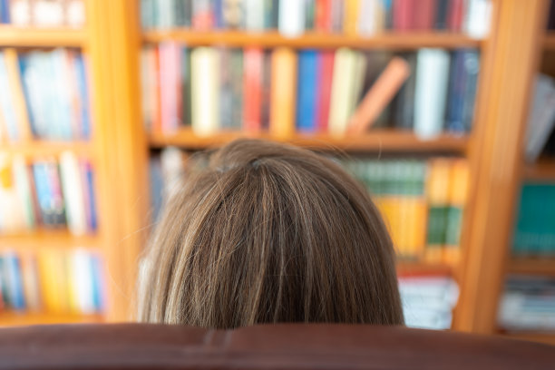 一个背靠椅背坐在扶手椅上的女人，面前放着一个装满书的大书柜。图片下载