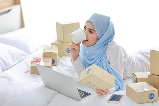 年轻迷人的亚洲女人穿着睡衣，穿着穆斯林服装，拿着咖啡杯躺在床上，用电脑和在线包裹箱送货。创业中小企业自由职业女孩用手机工作图片下载