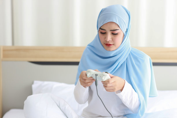 VDO游戏控制台概念。活跃的亚洲妇女穿着白色的穆斯林睡衣坐在床上，拿着操纵杆玩着令人兴奋的游戏。可爱的女孩拿着游戏控制器看起来很兴奋图片下载