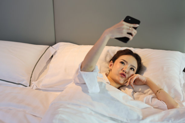 俯视图年轻美丽的亚洲女人穿着白色睡衣躺在床上自拍，在做好梦之前用手机打招呼。生活方式的概念图片下载