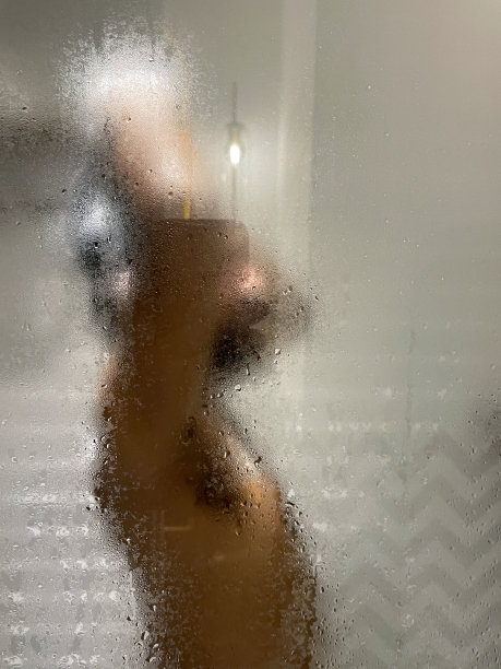 印度男子在浴室里洗头，身后是蒸汽蒸汽的玻璃淋浴间屏幕，让洗发水泡沫在流水下卷曲，白色斜边墙砖呈水平堆叠图案，侧视图，聚焦于前景图片下载