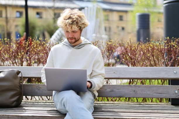 一个留着胡子的男人拿着笔记本电脑坐在公园的长椅上。图片下载