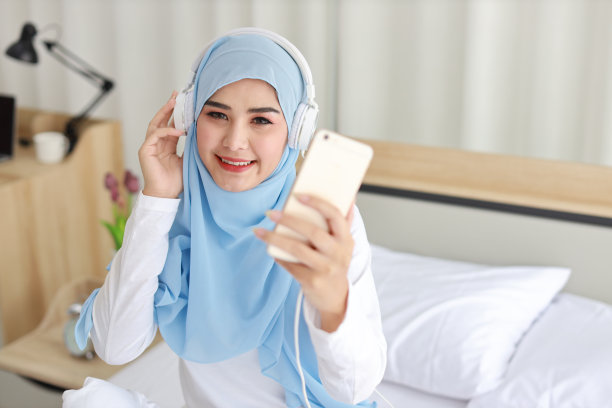 美丽的亚洲女性肖像，穿着穆斯林睡衣，在手机上观看网络故事，躺在床上，连接无线互联网。年轻可爱的女孩戴着头巾，用智能手机听音乐图片下载