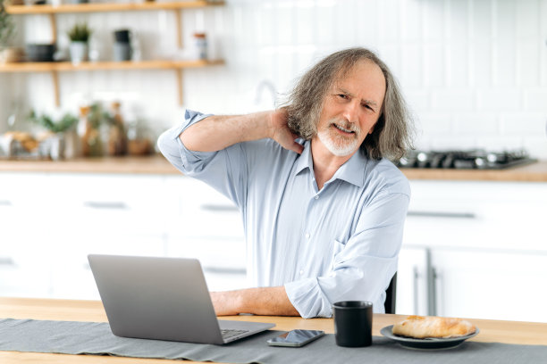 不健康的白头发老人，坐在厨房的桌子上，拿着笔记本电脑，感觉劳累过度，疲倦，脖子痛，按摩脖子，悲伤地看着镜头，做着鬼脸图片下载