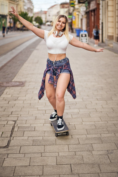 快乐的少女在城市街道上玩滑板，同时听着耳机上的音乐-保持微笑，享受生活的概念-背光夏日生活方式照片图片下载