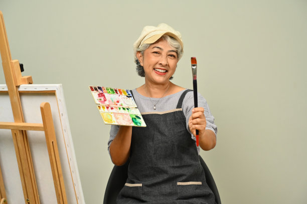 面带微笑的白发老妇人拿着调色板和画笔坐在米色背景的画架前。人，闲和爱好。图片下载