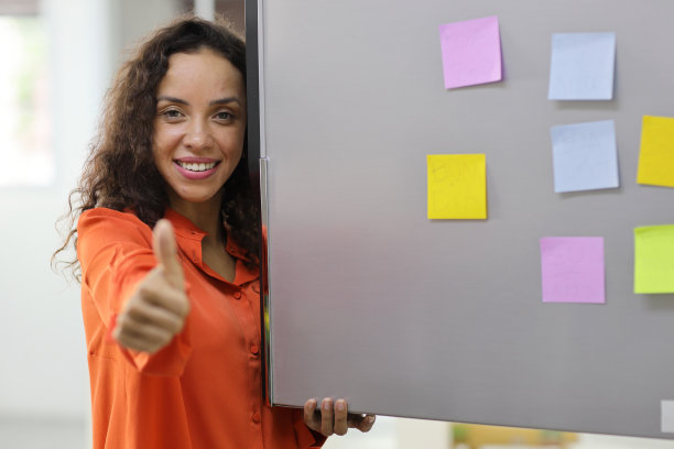 聪明而活跃的拉丁女人站在那里，拿着不同颜色的笔记纸，准备在厨房的冰箱上留言。女性竖起大拇指，微笑着盯着镜头。图片下载