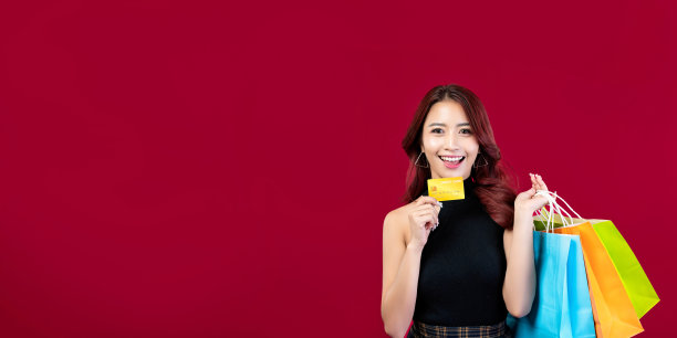 横幅:时尚的亚洲女人穿着黑色连衣裙，拿着购物袋，拿着深红色背景的金色信用卡。漂亮的泰国女人拿着购物袋拿着红色背景的金色信用卡图片下载