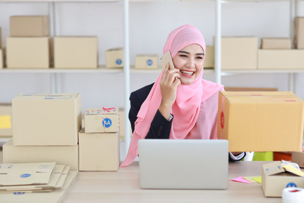 身着穆斯林服装的虔诚的亚洲妇女坐在那里，用智能手机和顾客交谈，脸上带着快乐的表情和商业包裹。图片下载