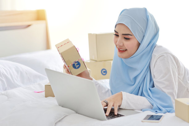 美丽和年轻的亚洲妇女穿着穆斯林睡衣，有吸引力的样子，躺在床上的电脑和在线包裹盒交付。创业中小企业自由职业女孩用手机工作图片下载