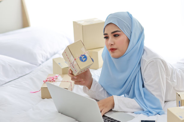 美丽和年轻的亚洲妇女穿着穆斯林睡衣，有吸引力的样子，躺在床上的电脑和在线包裹盒交付。创业中小企业自由职业女孩用手机工作图片下载