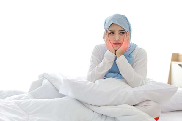 美丽和年轻的亚洲穆斯林妇女穿着睡衣与牙齿疼痛情绪坐在床上。可爱的女孩用手抚摸脸颊，牙疼得难受。牙齿保健和健康的理念。图片下载