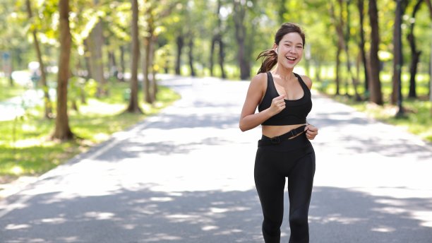 美丽的亚洲健身女运动员在户外进行晨跑锻炼训练。快乐的微笑运动员女子在公园的树下跑步，同时看着相机和听音乐图片下载