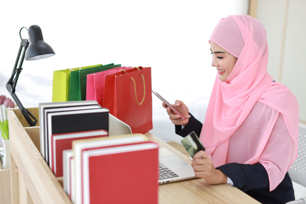 活跃的聪明的亚洲妇女穿着蓝色的穆斯林西装和粉红色的轴坐着，拿着信用卡，同时使用手机支付或在线购物。创业小生意的女孩在家工作。图片下载