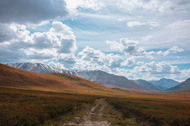 五彩缤纷的秋季景观，高原上的丘陵和阳光普照的雪山山脉在戏剧性的多云天空下。鲜艳的秋色笼罩着群山。多变天气里的阳光和云影。图片下载