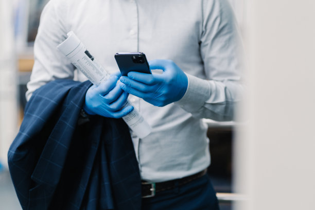 穿着优雅服装的陌生商人在现代智能手机上发信息，在大流行期间戴着一次性防护手套，拿着卷起的报纸和黑色夹克，在网上看新闻图片下载