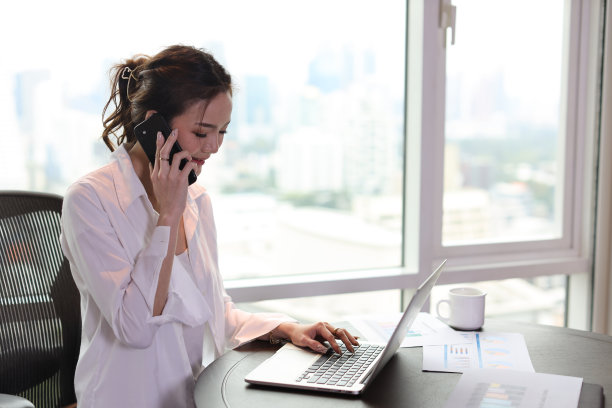 自信和美丽的年轻亚洲女商人的肖像，带着快乐的脸，坐在与城市背景的酒店使用电脑和手机。成功的商业和生活理念图片下载