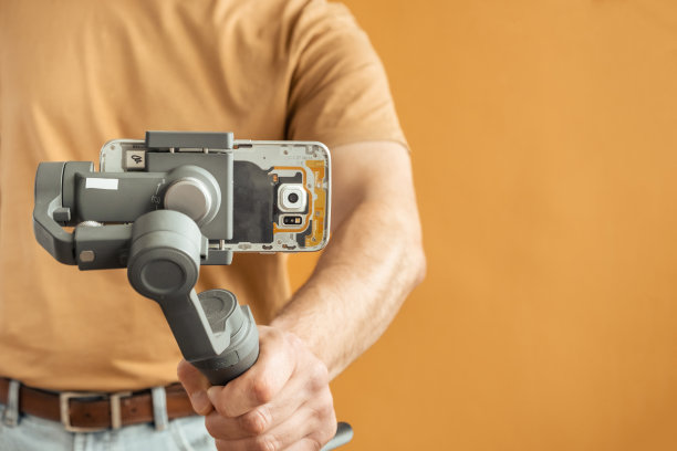 一名男子用智能手机在黄色背景上使用万向节稳定器进行拍摄。使用三轴电子稳定器进行vlog或视频拍摄。图片下载