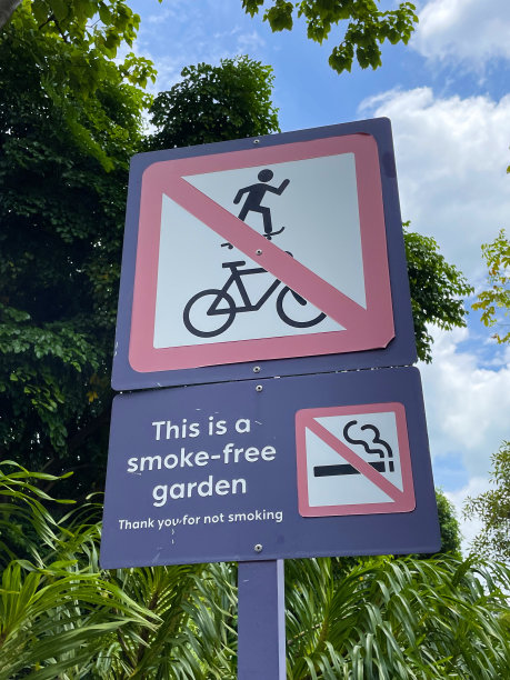 公园内禁止骑自行车、滑板或吸烟的标志，在杆子、树冠和多云、蓝色晴朗的天空背景上，特写图像，重点放在前景上图片下载