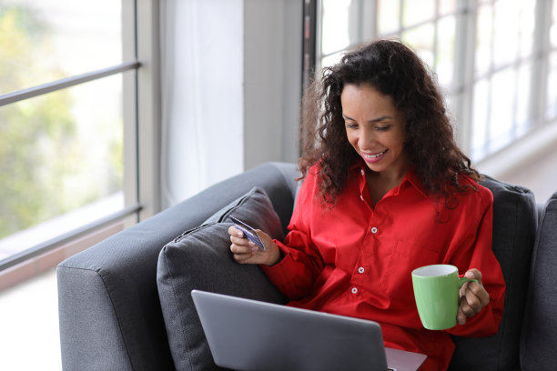 快乐微笑的拉丁女人坐在沙发上喝咖啡，在客厅用智能手机上网。科技生活方式图片下载