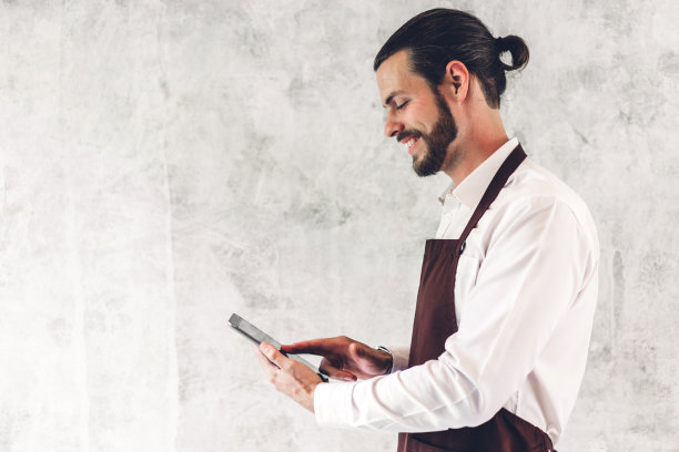 蓄着大胡子的帅气咖啡师和小企业主的肖像，微笑着拿着平板电脑在咖啡馆的墙壁背景上图片下载