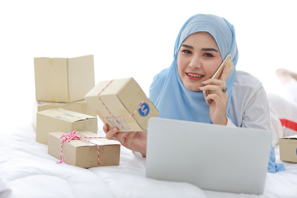 美丽和年轻的亚洲妇女穿着穆斯林睡衣，有吸引力的样子，躺在床上的电脑和在线包裹盒交付。创业中小企业自由职业女孩用手机工作图片下载