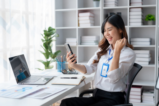 快乐的亚洲女商人举起胜利的手，拿着智能手机开心地微笑。工作中成功的概念。图片下载
