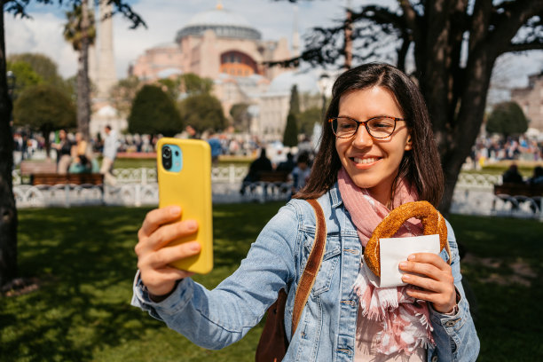 一名年轻女子在伊斯坦布尔圣索菲亚大教堂前边吃椒盐卷饼边自拍图片下载