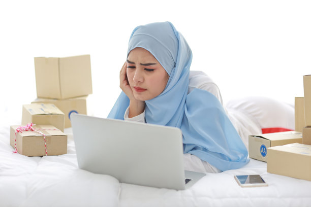 年轻漂亮的亚洲女人穿着穆斯林睡衣，带着头痛的情绪躺在床上，电脑、手机和在线快递。戴头巾的聪明女孩收到了坏消息，很累图片下载