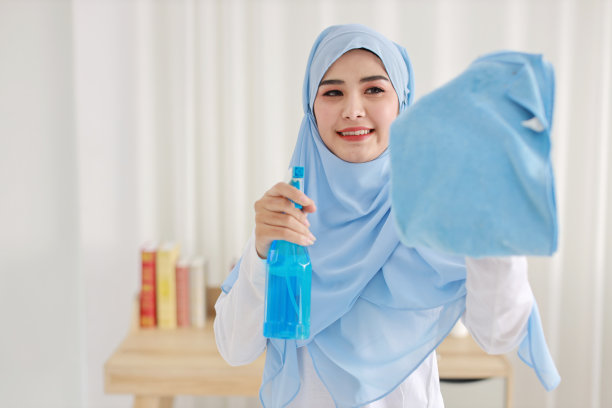 室内镜头活跃的年轻美丽的亚洲穆斯林家庭主妇手持清洁喷雾器，清洗窗户玻璃。可爱的女孩在搬进新公寓后快乐地打扫卧室。图片下载