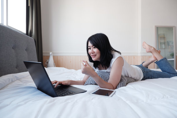 亚洲年轻女子躺在卧室的白色床上，在笔记本上写字。女商人用笔记本电脑和技术概念在家工作。图片下载