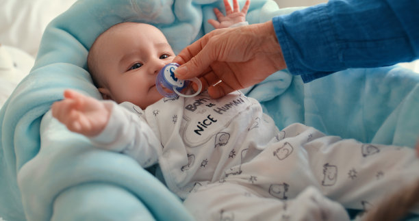 新生儿，婴儿安抚奶嘴和男孩与母亲的手安慰，安全和保障。可爱和健康的婴儿孩子与一个假人的发展，成长和照顾在一个毯子在家里图片下载