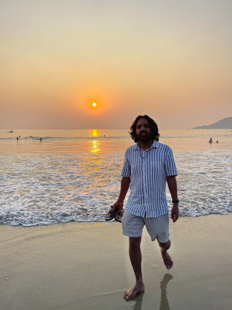 印度果阿，帕洛莱姆，一个穿着短裤和条纹短袖衬衫的印度男人，在沙滩上拿着凉鞋的特写照片，黄昏时分，太阳落山，度假游客赤脚走在海边，焦点在前景图片下载