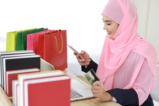 活跃的聪明的亚洲妇女穿着蓝色的穆斯林西装和粉红色的轴坐着，拿着信用卡，同时使用手机支付或在线购物。创业小生意的女孩在家工作。图片下载