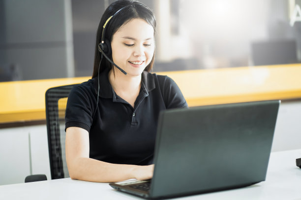 友好的亚洲女警官戴着耳机在呼叫中心的电脑前愉快地微笑着。图片下载