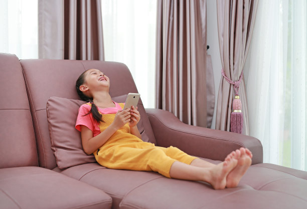 亚洲小女孩在家里客厅的沙发上玩智能手机时大笑的照片图片下载