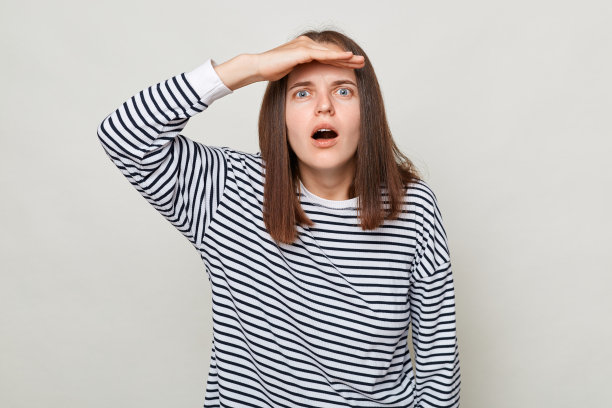 震惊惊讶的棕色头发的女人，穿着条纹衬衫，孤立地站在灰色的背景上，用手靠近额头，看着大大的眼睛。图片下载