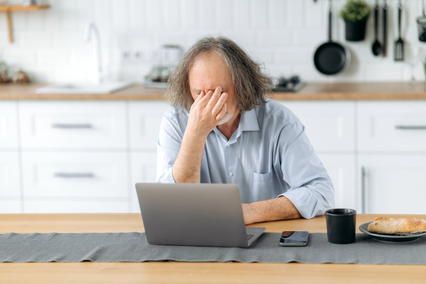 疲劳的概念。精疲力竭的白头发老人，退休老人，坐在厨房的桌子旁，手里拿着笔记本电脑，感觉工作过度，压力很大，用笔记本电脑工作很疲劳，有偏头痛，需要休息图片下载