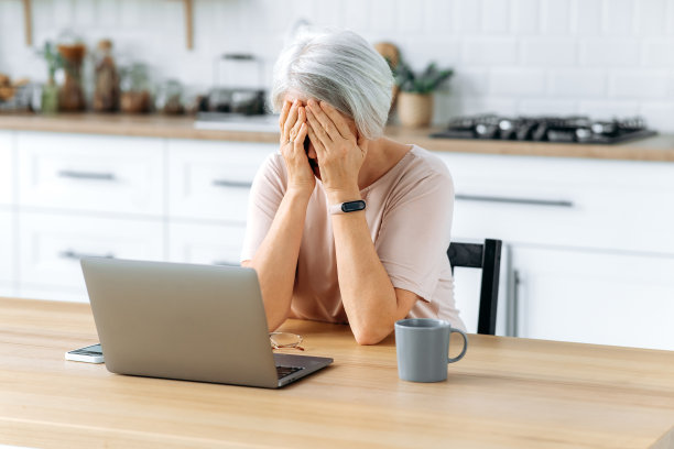 疲劳的概念。精疲力竭的中年白头发白人妇女，退休人员，坐在厨房的桌子上，用笔记本电脑工作，感到过度劳累和压力，因为使用笔记本电脑而感到疲劳，患有偏头痛图片下载