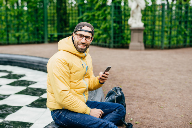 大胡子的时髦男性的横向肖像，带着愉快的表情看着旁边，坐在喷泉旁拍摄美丽的大自然。有型的男人拿着手机在户外休息图片下载