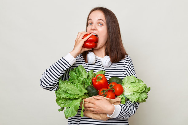 饥饿的棕色头发的女人，穿着条纹衬衫，站在灰色的背景中，拿着新鲜的时令蔬菜，咬着甜椒。图片下载