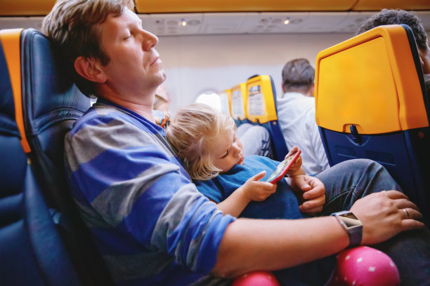 年轻的父亲抱着蹒跚学步的女儿在飞机上度假。疲惫的男人和可爱的女孩在夜间飞行。孩子在玩智能手机。一家人坐在飞机上图片下载