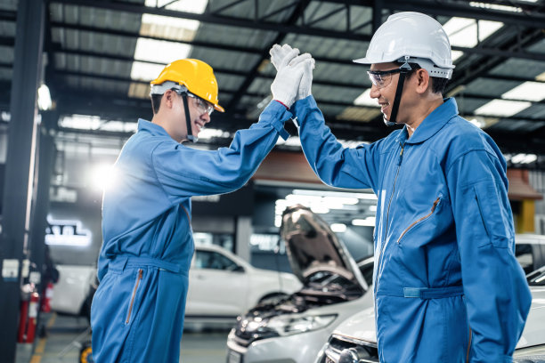 亚洲两名汽车技工戴着头盔在机械厂工作。在机修厂车间，发动机机器修理成功后，汽车维修工团队高兴地握手。图片下载