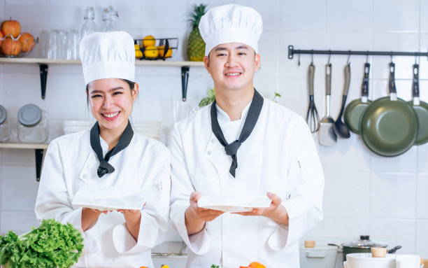 两位亚洲职业厨师夫妇身穿白色制服，头戴帽子，露出空白的盘子，大拇指向上，一起在厨房里做饭，微笑着幸福，充满信心。食品的概念。图片下载