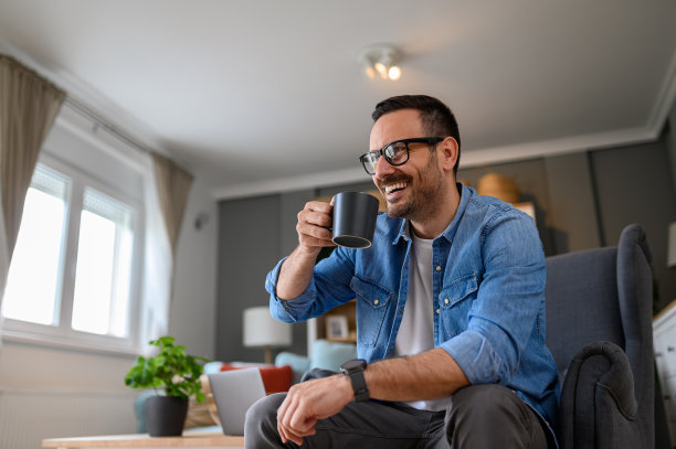 年轻商人坐在扶手椅上愉快地笑着喝咖啡的低角度视图。快乐的男性自由职业者在他的咖啡休息时间在家办公室里思考新的想法图片下载